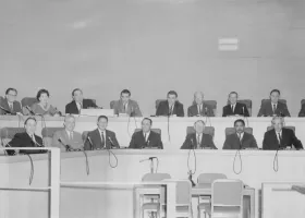 A black-and-white photo shows a group of people seated in a formal setting, likely a conference room or council chamber. They are arranged in two rows behind microphones on desks. The room has a modern design with clean lines and minimal decor.