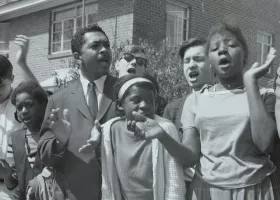 A group of young people and a man in a suit stand together outside, clapping and singing with expressive faces in front of a brick building, appearing to participate in a civil rights demonstration.