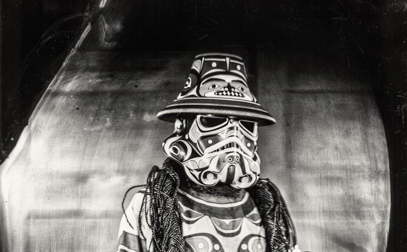 A person dressed in an intricately designed costume featuring Native American patterns and symbols, including a mask resembling a Stormtrooper helmet and a wide-brimmed hat. They hold a small decorated object in one hand and wear multiple necklaces. Black and white photo.