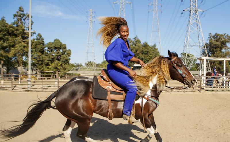 A person wearing a bright blue outfit rides a galloping horse in a fenced sandy arena. The rider's hair flows back dramatically. Power lines and towers are visible in the background under a clear blue sky.