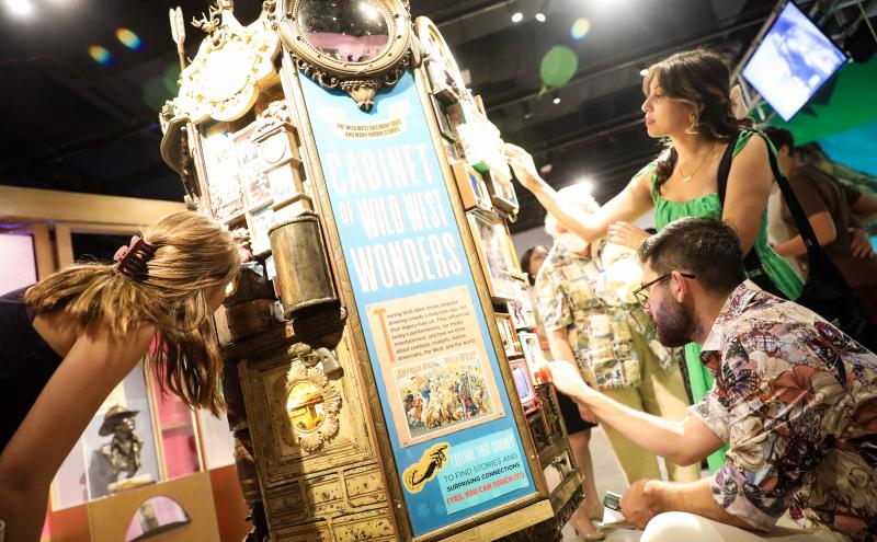 Three adults examine an ornate exhibit titled "Cabinet of Wild West Wonders" in a brightly lit museum, engaging with its various interactive elements and displays.