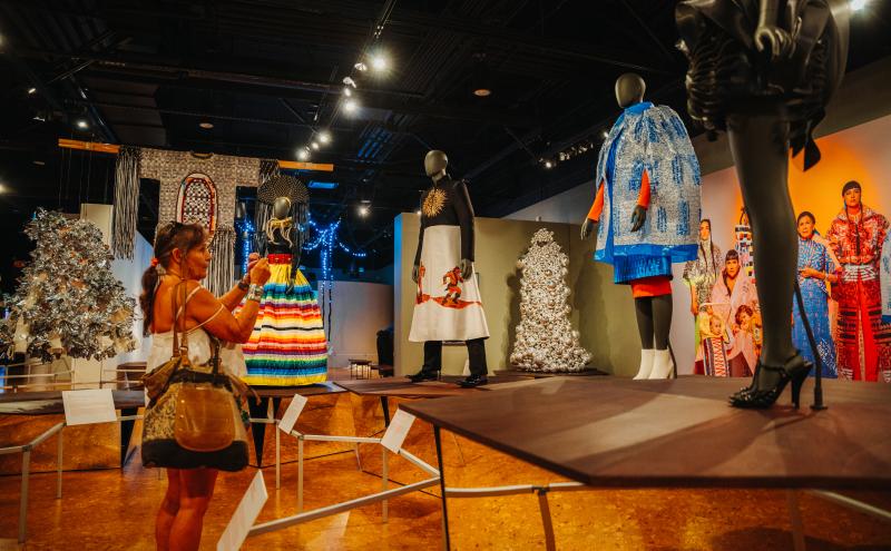 A woman takes a photo of mannequins dressed in colorful, elaborate outfits on display in a museum exhibit with dramatic lighting and various artwork in the background.