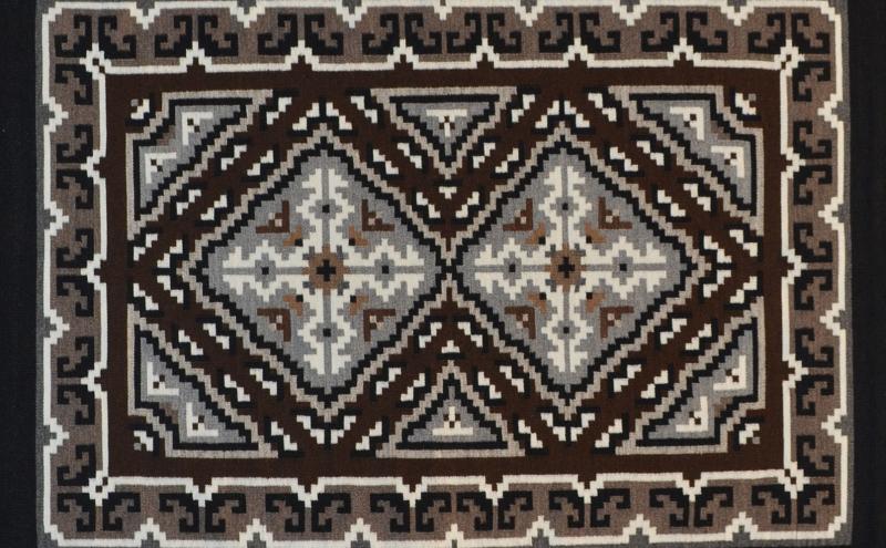 A rectangular rug with a geometric pattern in black, white, gray, and brown tones, featuring symmetrical diamond shapes and angular borders.