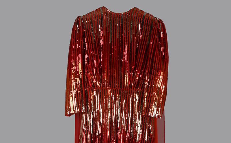 A long, ankle-length red dress with three-quarter sleeves, covered in shiny sequins arranged in vertical lines, displayed against a plain gray background.