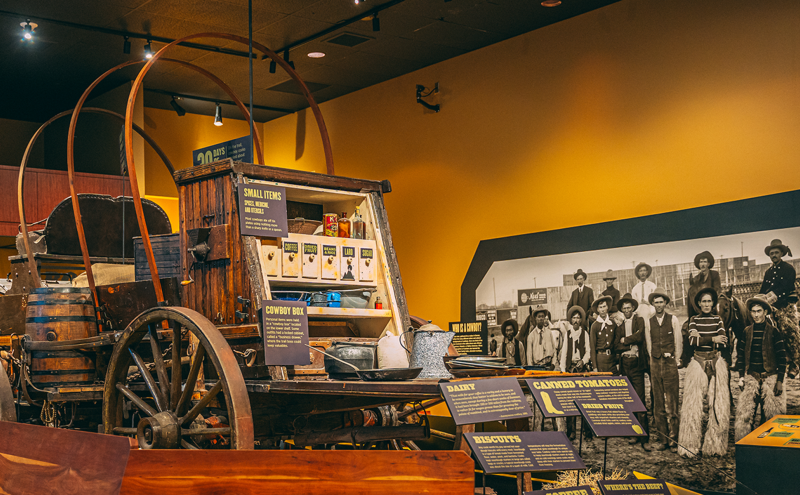 A museum exhibit features a wooden covered wagon stocked with supplies, surrounded by informational signs. Behind it, a large black-and-white photo shows a group of people in period Western clothing.