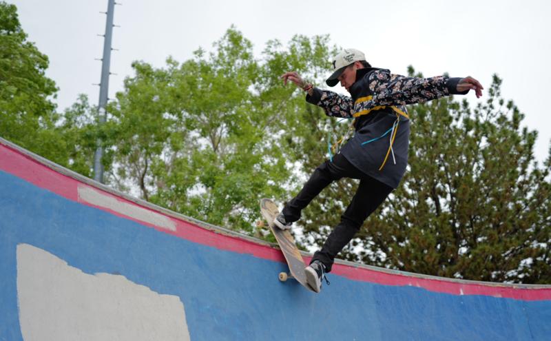 A skateboarder wearing a patterned shirt, black pants, and a white helmet performs a trick on the curved edge of a colorful skatepark ramp, with trees and a cloudy sky in the background.