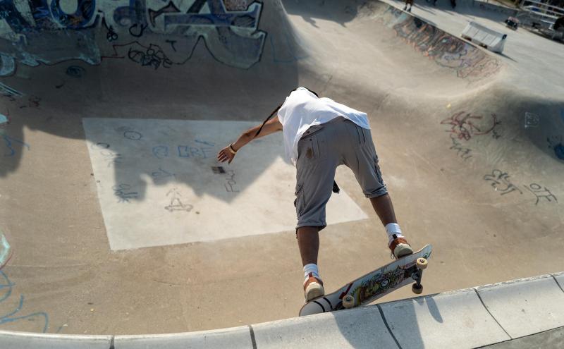 A skateboarder in a white shirt and gray shorts prepares to drop into a concrete skatepark bowl covered in graffiti, balancing on the edge with knees bent and arms outstretched.