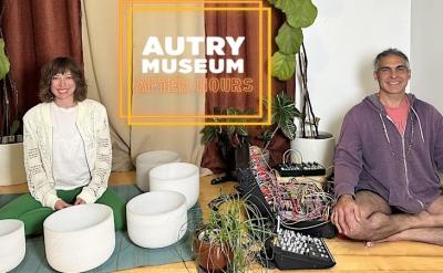 Two people are sitting on the floor cross-legged, surrounded by various instruments and plants. A sign reads "Autry Museum After Hours." They appear relaxed, with one person wearing a white sweater and green pants, and the other in a purple hoodie and shorts.