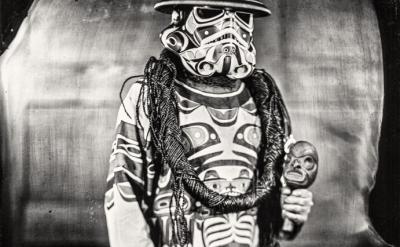 A person is dressed in a Star Wars stormtrooper helmet painted in Indigenous art style, wearing a matching robe, and holding a small carved mask. The background is blurred and the image has a vintage, black and white aesthetic.