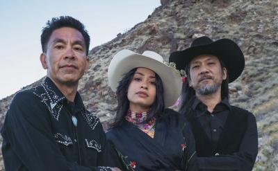  Photo of The Lion Heart. Three people stand in front of a rocky hillside. The person on the left wears a black shirt with white embroidery, the person in the middle wears a white cowboy hat and a black dress, and the person on the right wears a black cowboy hat and a sleeveless vest.