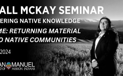 Black and white image of a person standing in a field of tall grass with hills and a cloudy sky in the background. Text reads, "Marshall McKay Seminar for empowering native knowledge. Going Home: Returning Material Culture to Native Communities. November 15-16, 2024." Presented by San Manuel Band of Mission Indians.