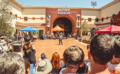 People gathered outdoors watching a performer at the Autry Museum. The museum's entrance is in the background, with banners on either side and tents providing shade over some spectators. The scene is lively with a mix of standing and seated onlookers.