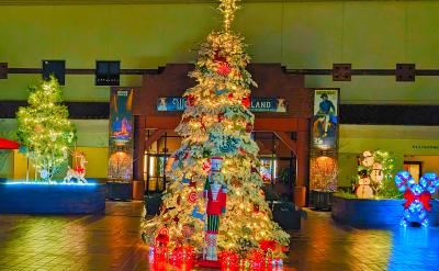 A festive holiday display featuring a large decorated Christmas tree adorned with lights and ornaments, with a nutcracker figure in front. The backdrop includes a building entrance with additional lit decorations, banners, and a snowman made of lights.