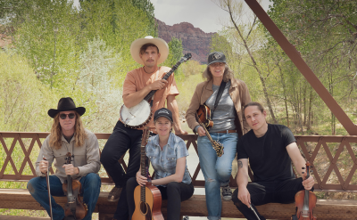 Five people pose with musical instruments on a wooden bridge surrounded by greenery and mountains. From left to right: they hold a violin, banjo, acoustic guitar, mandolin, and another violin. Some wear hats and casual attire, and they appear relaxed and smiling.