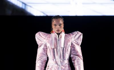 A model walks down a lit runway wearing a metallic pink, futuristic, padded jumpsuit with exaggerated shoulders. Her hair is styled in braids, and she pairs the outfit with white platform shoes and red statement earrings. The background is dark, highlighting the runway.