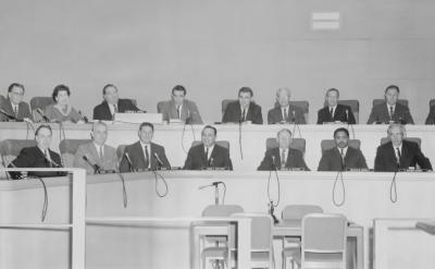 A black-and-white photo shows a group of people seated in a formal setting, likely a conference room or council chamber. They are arranged in two rows behind microphones on desks. The room has a modern design with clean lines and minimal decor.