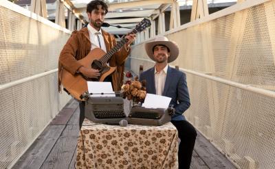 Two men standing on an outdoor bridge with a netted railing. One stands playing an acoustic guitar, wearing a brown suede jacket with fringe. The other sits at a table covered with a floral cloth, wearing a white hat and suit, with two typewriters in front of him.