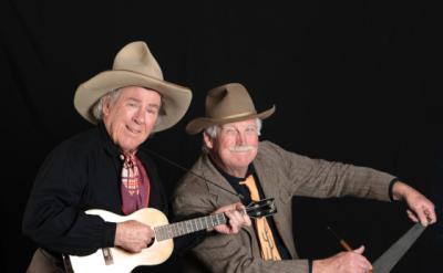 Two elderly men in cowboy hats smiling. One man is holding a small guitar, possibly a ukulele, and the other is playing a large hand saw with a bow. They are dressed in western-style clothing and are posing against a plain black background.