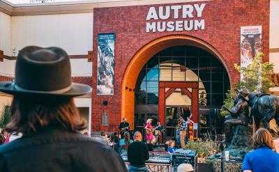 Outdoor concert at the Autry Museum with musicians performing on stage. A crowd watches, including a person in the foreground wearing a wide-brimmed hat. Brick building and statues enhance the vibrant scene.