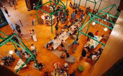 A busy indoor event with people gathered around tables covered in assorted materials. Green metal frames hang above, and various activities are happening throughout the space. Attendees include families and individuals in a creative workshop setting.