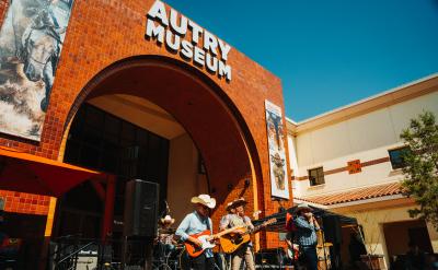 A country band performs outside the Autry Museum under a clear blue sky. Musicians play guitars and a drum set. The museum building features a red brick facade and large entrance arch. Banners with Western art hang on display.