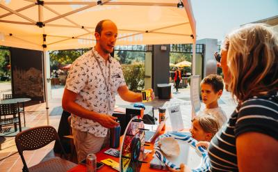 A man stands at a booth under an orange canopy, interacting with two children and a woman holding a paper plate. The booth has a colorful wheel and various items on a table, suggesting a fun, outdoor event.