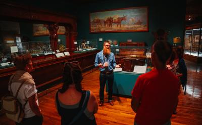 A guide in a denim shirt addresses a small group of people in a dimly lit museum room, surrounded by exhibits and artifacts. A large painting of animals hangs on the wall behind them.