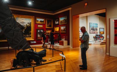 A man stands in an art gallery observing a collection of paintings on red walls. Two other people are viewing exhibits, including a large sculpture and a motorcycle displayed nearby. The room is warmly lit with wooden flooring.