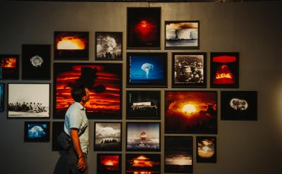 A person observes a gallery wall displaying various framed photographs of nuclear explosions. The images depict dramatic mushroom clouds and fiery skies, capturing the powerful and destructive force of the blasts.