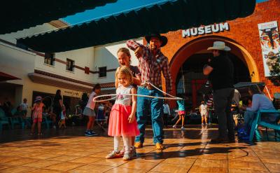 A man helps a young girl hula hoop on a sunlit patio outside a museum. They're surrounded by people, some in cowboy hats. The building in the background has a brick façade with "MUSEUM" written above the entrance.