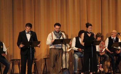 A group of people stand on stage in front of music stands, reading from scripts. They appear to be performing or rehearsing, with some seated in the background. The stage is set against a backdrop of long curtains.