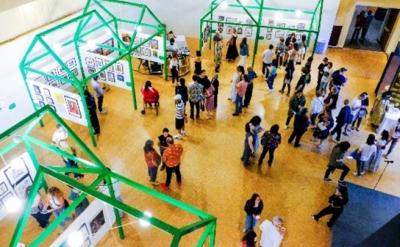 Aerial view of an art gallery with green geometric structures. People browse and converse, examining artworks displayed on the walls. The setting is bright and spacious, creating a lively atmosphere.