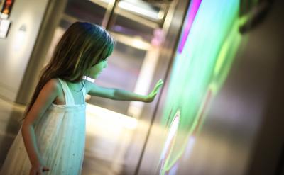 A young girl in a white dress reaches towards a brightly illuminated, colorful wall display in a dimly lit room, her face glowing with light from the screen.