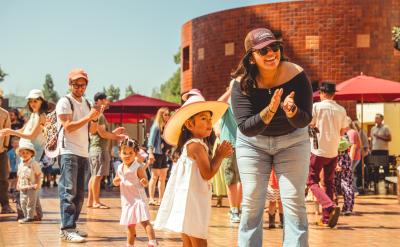 A joyful outdoor scene featuring a group of people dancing and clapping under a sunny sky. A woman wearing sunglasses and a hat claps alongside a young girl in a pink cowboy hat. Other adults and children join the lively gathering.