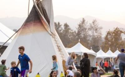 People enjoying an outdoor event with tents and teepees. Adults and children are walking and playing, surrounded by trees and mountains under a hazy sky.