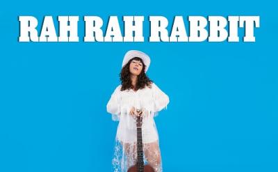 A woman in a white dress and hat stands against a bright blue background, holding a guitar vertically. The text "Rah Rah Rabbit Ghosts" is displayed prominently at the top and bottom of the image.