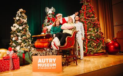 A group of four people pose in a festive setting at the Autry Museum. Santa sits in a sleigh surrounded by decorated Christmas trees and wrapped gifts. A sign reads "Autry Museum of the American West" in front of them.