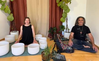 Two people seated on a wooden floor surrounded by sound bowls and electronic equipment. The background features tall green plants and curtains, creating a serene atmosphere. Both individuals are smiling.