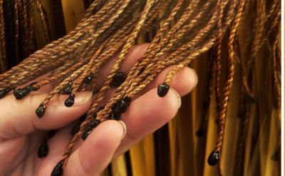 A hand holding a bunch of brown, twisted cords with black tips. On the right, a shiny, decorative object with a wavy, iridescent design is attached to similar cords. The background features more cords hanging vertically.