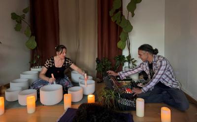 A woman and man seated on the floor playing white crystal singing bowls and electronic synthesizers in a dimly lit room with plants and candles.