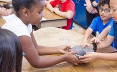 Children sitting at a classroom table, engaging with a large rock. Two children in the foreground hold the rock together, while others watch. The group is focused and appears to be participating in a hands-on activity.