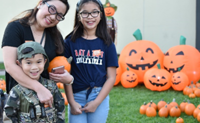 A woman and two children smile in front of Halloween decorations, including large inflatable pumpkins with jack-o'-lantern faces. The boy wears a camouflage outfit, and the girl wears glasses and a casual outfit. A scarecrow stands in the background.