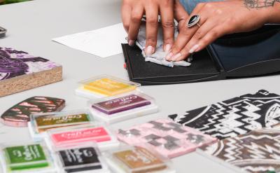 A person with tattoos on their left arm presses a stamp onto an ink pad at a table. Various colorful ink pads and patterned stamps are arranged around them. A lush, green background is visible in the distance.