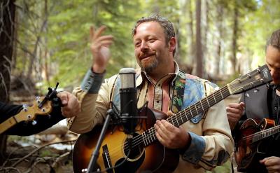 A man with a guitar stands in a forest, smiling as he performs. He is surrounded by two other musicians, one holding a guitar and the other playing a mandolin. A microphone is positioned in front of them. Trees and greenery fill the background.