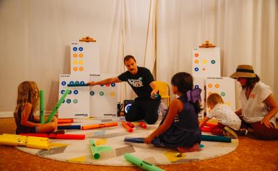 A man sits on the floor with a group of children, holding colorful foam tubes. Charts with symbols are in the background. The children, wearing casual clothes, listen attentively as they interact with the materials in a well-lit room.