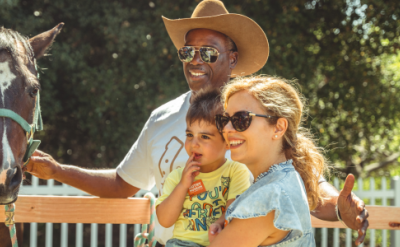 A man wearing a cowboy hat and sunglasses stands with a woman and a young child in front of a horse. The woman holds the child, who holds a snack. They are outside near a wooden fence and greenery.