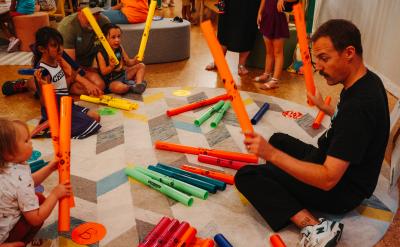 A group of children and adults gather in a colorful play area, engaging with bright music tubes and instruments. They're seated on a patterned rug, actively exploring sounds and rhythms together, creating a lively and interactive atmosphere.