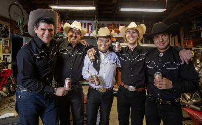 Five men wearing cowboy hats and black shirts with white trim stand in a garage, smiling and holding beer cans. They are surrounded by various tools and equipment, and a motorcycle is partially visible in the background.