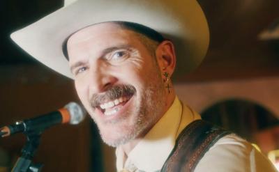 A person wearing a cowboy hat and earring smiles while singing into a microphone. The background is dimly lit, creating a warm ambiance.