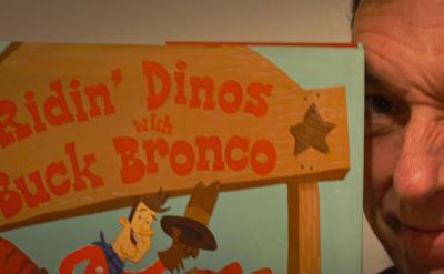 A person winking while holding a book titled "Ridin' Dinos with Buck Bronco" and a blue puppet. The puppet has large eyes and appears to be interacting with the person.